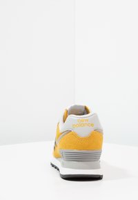 Gula och grå New Balance-sneakers sedda bakifrån, som visar det texturerade mockaskinnet och den grå sulan på en vit yta.