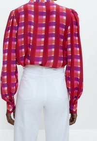 Blusa a quadri rossi e rosa con maniche lunghe e voluminose e polsini con bottoni, abbinata a pantaloni bianchi a vita alta, che presentano una texture liscia.
