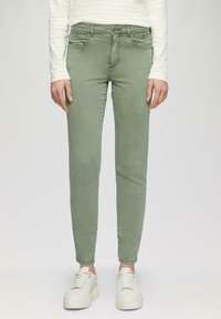 Zielone dżinsy skinny o gładkiej fakturze, z przednimi kieszeniami i pojedynczym zapięciem na guzik, w zestawie z białymi trampkami.