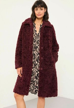 Burgunderfarbener Kunstpelzmantel mit Kragen, langen Ärmeln und vorderen Taschen, mit Knopfverschlüssen. Getragen über einem schwarz-weißen gemusterten Kleid.