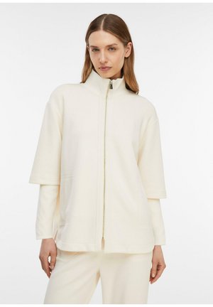 Frau trägt eine cremefarbene, kurzärmelige Reißverschlussjacke über einem langärmeligen Shirt, dazu passende cremefarbene Hosen, steht vor weißem Hintergrund.