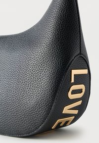 Κοντινό πλάνο γωνίας μαύρης δερμάτινης τσάντας με υφή, με χρυσά μεταλλικά γράμματα που σχηματίζουν τη λέξη "ΑΓΑΠΗ" κάθετα στη πλευρά.