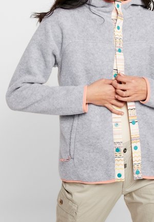 Manos abrochando botones en una chaqueta de forro polar gris claro con ribete en tonos pastel, llevada sobre pantalones cargo beige.