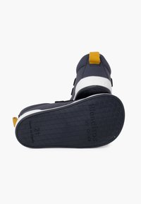 Námořnická obuv s žlutým akcentem na patě. Má bílou podrážku s texturovaným gripem. Design zahrnuje nastavitelný pásek a polstrovaný svršek.