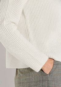 Persoon in een witte gebreide trui met de hand in de zak van grijs geruite broeken, waarbij textuur en stofdetails worden getoond.