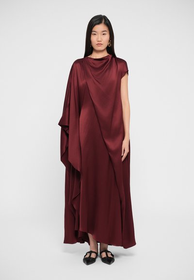 Lang, flydende kjole i dyb bordeaux satin. Funktioner draperet design med asymmetrisk kant og korte ærmer, parret med sorte flade sko.