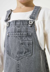 Grijze denim overall met metalen clipsluitingen, voorzak en gestructureerde stof. gedragen over een witte top met lange mouwen en gathered mouwen.