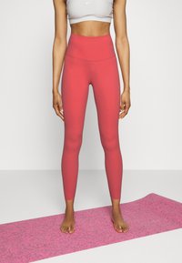 Leggings rouges taille haute en tissu extensible, avec une texture lisse et un détail de logo discret, associés à un tapis de yoga rose.