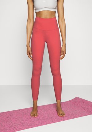 Leggings rouges taille haute en tissu extensible, avec une texture lisse et un détail de logo discret, associés à un tapis de yoga rose.