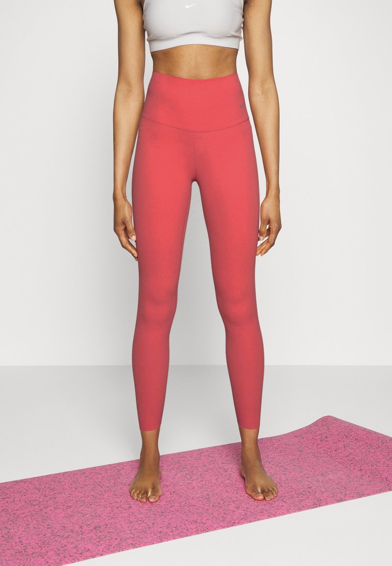 Leggings rouges taille haute en tissu extensible, avec une texture lisse et un détail de logo discret, associés à un tapis de yoga rose.