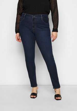Vrouw draagt donkerblauwe skinny jeans, zwarte doorschijnende blouse met polkadots en zwarte open hak sandalen, staand tegen een effen achtergrond.