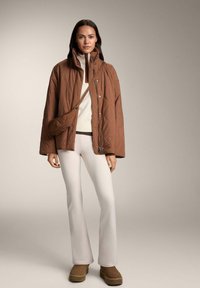 Smeđa prošivena jakna s kopčama na drukere, u kombinaciji s krem džemperom i svijetlim hlačama. Ukrašena malom smeđom torbicom preko ramena.