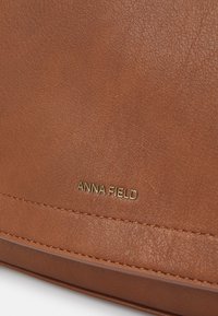 Bruin gestructureerd leren oppervlak met het geperste "ANNA FIELD" logo in goud, met een gestikte naad aan de onderkant.