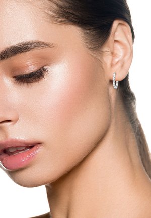 Primo piano del viso di una donna girato di lato, che mostra una pelle liscia, occhi chiusi con lunghe ciglia, labbra lucide e un orecchino ad anello argentato.