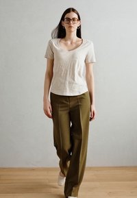 Tricou bej deschis cu decolteu în V, pantaloni largi verzi măslinii, model purtând ochelari, podea din lemn, fundal neutru.