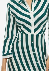 Robe chemise à rayures vertes et blanches avec un devant boutonné, un col et des manches roulées. Le tissu a une texture lisse et un design épuré.