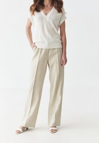 Bílý pletený svetr s výstřihem do V, volný střih; béžové lněné kalhoty širokých nohavic; bílé nazouvací sandály; minimalistický design, hladká textura.