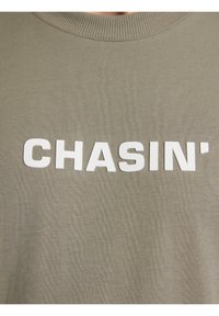 Olajzöld pulóver, bordás kerek nyakkal és kiemelkedő fehér felirattal "CHASIN'" az elején. Puha tapintás, laza kialakítás.