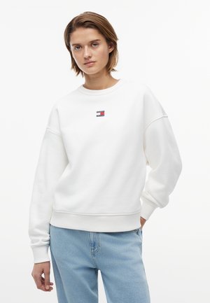 Νεαρό άτομο με κοντό καστανό μαλλί, φορώντας άσπρο φούτερ Tommy Jeans και ανοιχτό μπλε τζιν, σε απλό λευκό φόντο.