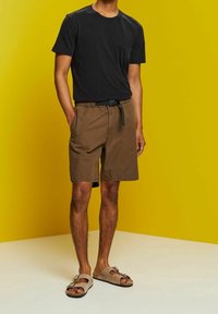 Homme portant un t-shirt noir, un short marron avec ceinture et des sandales beiges, debout devant un fond jaune les mains dans les poches.