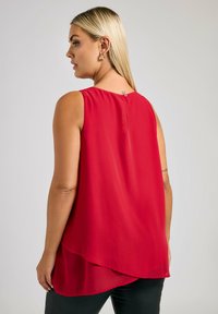 Röd ärmlös blus med hög-låg fåll och knappstängning på baksidan. Tillverkad av lätt, mjukt tyg med en flödande design.