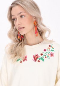 Krém színű pulóver színes virágmintával, rózsaszín, piros és zöld hímzéssel. A modellen hosszú fülbevalók vannak narancssárga és rózsaszín díszítéssel.