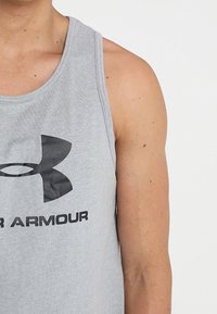Camiseta sin mangas gris de algodón con un escote redondo y un gran logo negro de Under Armour impreso en el frente.