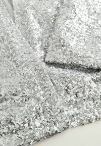 Silberner Paillettenstoff mit glänzender, strukturierter Oberfläche. Verfügt über dicht aneinander gereihte, kreisförmige Pailletten für ein schimmerndes Aussehen.