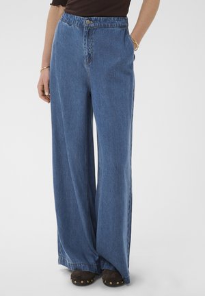 Femme portant un jean en denim bleu taille haute à jambes larges, des chaussures marron foncé avec des clous dorés, un haut noir et des bracelets dorés aux mains.