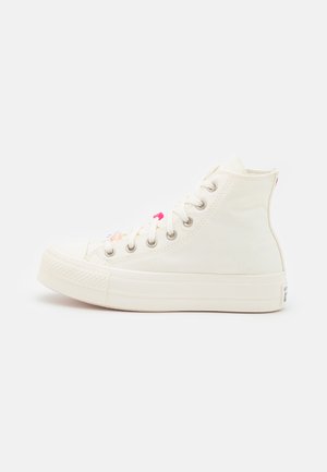 Zapatillas altas hechas de lona de color blanco roto con una suela de goma. Presentan acentos en rosa y ojales de metal plateado a lo largo del cordón.