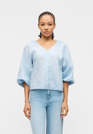 Junge Frau mit geflochtenem Haar trägt eine hellblaue bestickte Bluse mit Ballonärmeln und hell gewaschene Jeans, steht vor weißem Hintergrund.