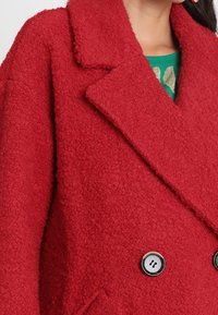 Rød uldfrakke med tekstur, udstyret med en reverskrave, dobbeltknappet design og to knapper. Har en hyggelig, blød finish.