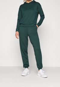Dark green hoodie and matching joggers, made from soft fabric, featuring a hood, elastic cuffs, and side pockets. Worn with white sneakers.