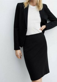 Blazer noir sur mesure porté sur un haut blanc ajusté, assorti à une jupe crayon noire. Tissu lisse avec un design structuré et des lignes simples.