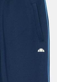 Pantalons de sport bleu marine avec une texture lisse, dotés de rayures latérales bleu clair et d'un logo blanc sur la partie inférieure de la poche.