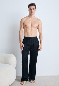 Black lightweight, loose-fitting track pants with an elastic waistband, smooth texture, and wide leg design, worn barefoot by a standing model.
