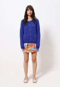 Femme debout contre un fond blanc portant un cardigan bleu vif à boutons, une jupe à motifs colorés et des ballerines beiges.