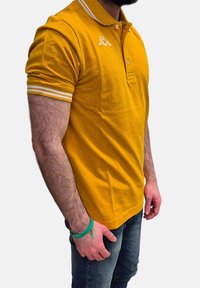 Polo giallo con tessuto strutturato, polsini a righe bianche e un piccolo logo sul petto. Abbinato a jeans blu.