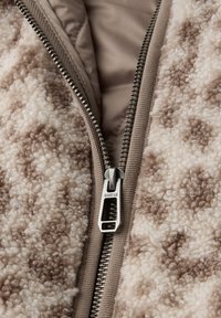 Gros plan d'une veste beige et marron partiellement zippée en tissu texturé laine, avec une fermeture éclair en métal et une doublure en tissu lisse.