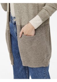 Cardigan aperto lungo in maglia beige-grigia, con tasca frontale e polsini a coste, indossato sopra un top crema testurizzato e jeans in denim.