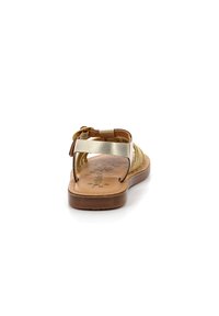 Sandale dorée et beige avec un design de sangle tressée, boucle ajustable et semelle plate. La semelle intérieure présente un motif texturé.