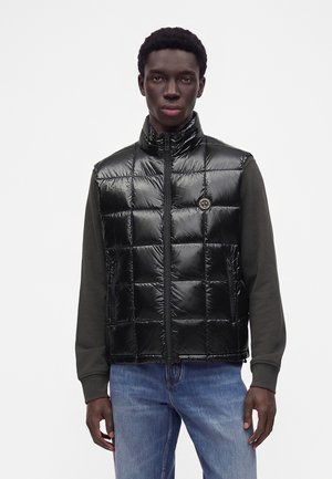 Man draagt een zwarte glanzende doorgestikte bodywarmer over een donker shirt met lange mouwen en een blauwe spijkerbroek, staand tegen een effen witte achtergrond.