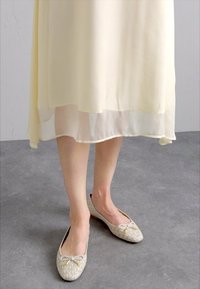 Ballerinas in hellem Beige mit einem dezenten Logomuster, runder Zehenpartie und kleinen Schleifendetails, kombiniert mit einem fließenden, blassgelben Kleid.