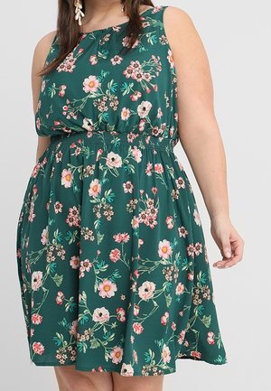 Ærmeløs grøn kjole med rosa og hvide blomsterprint, elastisk talje, båret af en person med langt hår og hængende øreringe.