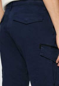 Granatowe spodnie cargo z wieloma kieszeniami, teksturowanym wykończeniem i tylną kieszenią z klapką. Materiał wydaje się być trwały i miękki.