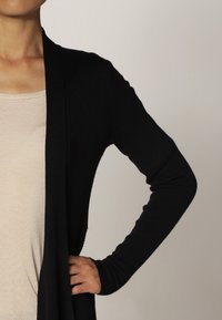 Svart ribbad cardigan med långa ärmar och öppen front, bärs över en ljusbeige åtsittande topp, visar en hand på höfterna.