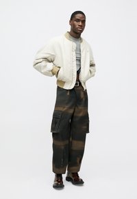 Beige bomber jacket with a ribbed collar, grey t-shirt underneath, and dark cargo pants featuring a brown tie-dye pattern. Brown boots.