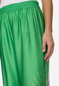 Zelené lehké kalhoty z látky s elastickým pasem, zdobené barevným květinovým potiskem na spodní části nohavic. Hladká textura a pohodlný volný střih.