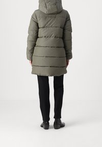 Chaqueta de plumas verde oliva con capucha, que presenta un acolchado horizontal, tejido texturizado y un corte de longitud media, combinada con pantalones y zapatos negros.