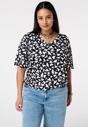 Vrouw met lang zwart haar, gekleed in een zwart-witte abstracte blouse met hartpatroon, blauwe jeans, gouden ketting en armbanden, staand.